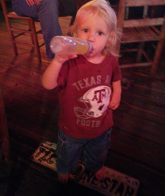 A baby with a bottle sanding on a hardwood floor that's patched with license plates