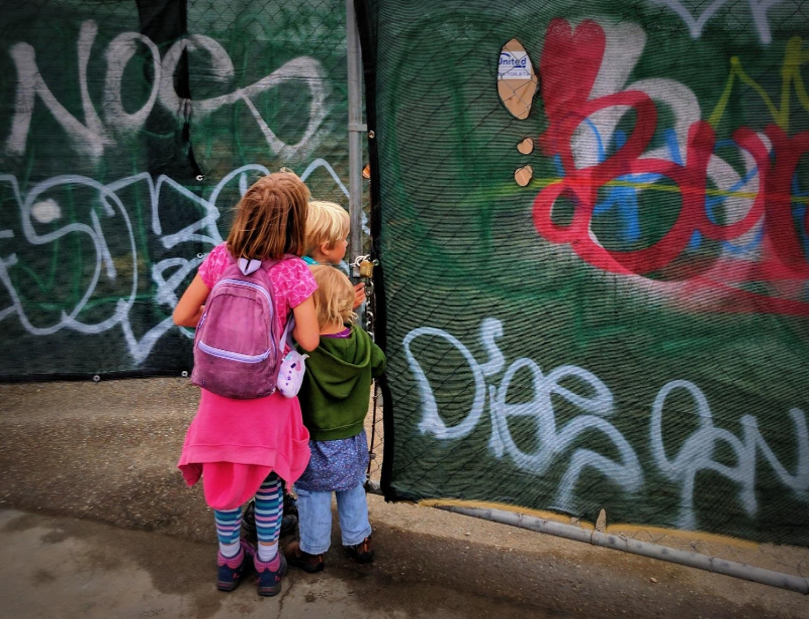 Three small kids peek around a fence with grafiti into a train yard 
                                                      construction project